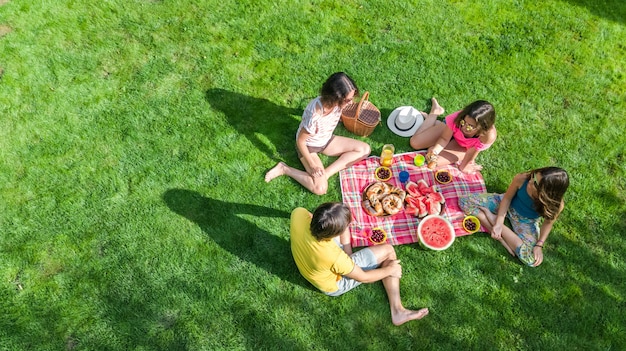 Happy family with children having picnic in park, parents with kids sitting on garden grass and eating healthy meals outdoors