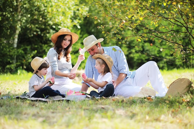 Famiglia felice con bambini che fanno picnic nel parco, genitori con bambini seduti sull'erba del giardino e che mangiano anguria all'aperto