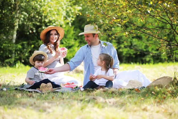 Famiglia felice con bambini che fanno picnic nel parco, genitori con bambini seduti sull'erba del giardino e che mangiano anguria all'aperto