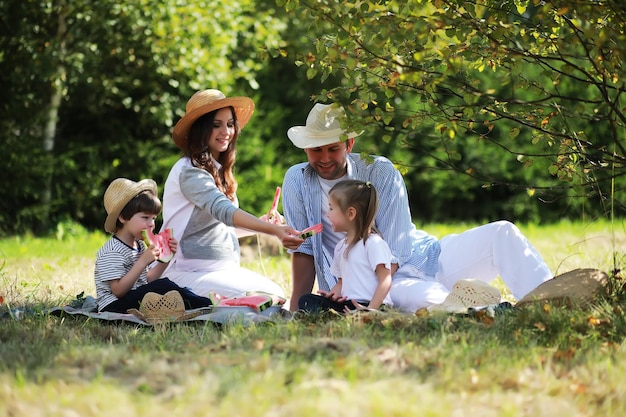 Happy family with children having picnic in park, parents with kids sitting on garden grass and eating watermelon outdoors