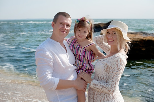 Счастливая семья с дочерью позирует возле моря, глядя в камеру