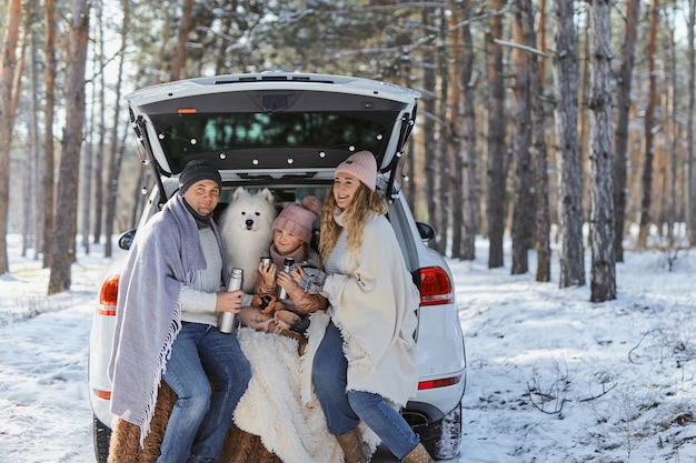 Happy family with dog on vacation during the winter holidays\
near road. dressed in warm clothes sitting on the trunk of a car\
and drinking tea from a thermos. space for text. winter\
vacation