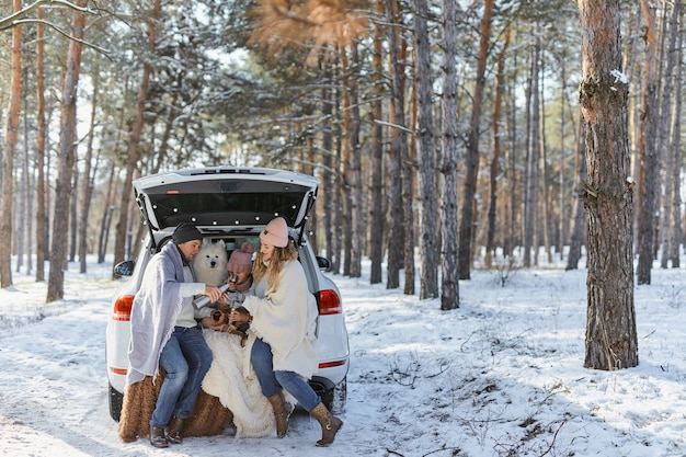 Happy family with dog on vacation during the winter holidays
near road. dressed in warm clothes sitting on the trunk of a car
and drinking tea from a thermos. space for text. winter
vacation