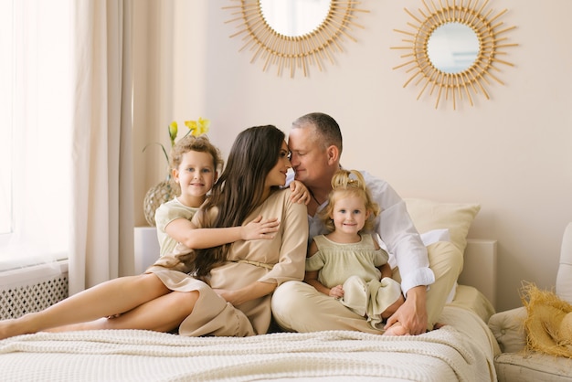 Happy family with little daughters sitting on the bed, mom and dad touched each other and smile, children hug their parents. Merry weekend morning