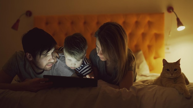 Happy family with little son and funny cat lying in bed at home and surfing social media