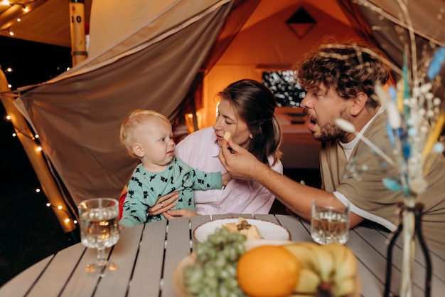 Happy family with lovely baby have dinner and spend time together in glamping on summer evening near cozy bonfire Luxury camping tent for outdoor recreation and recreation Lifestyle concept