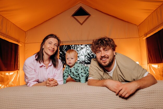 Happy family with lovely baby playing and spend time together in cozy glamping on summer evening while lying on bed Luxury camping tent for outdoor recreation and recreation