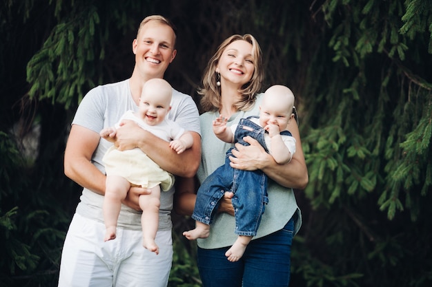 Happy family with twins boy and girl laughing posing together outdoors at green tree background. Smiling mother and father holding children enjoying parenthood