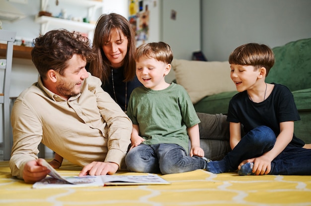 Счастливая семья с двумя маленькими сыновьями, читающими рассказ в помещении, родители с детьми проводят время вместе и лежат на полу дома