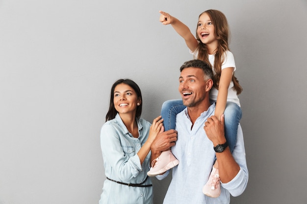 happy family woman and man smiling and looking aside while daughter sitting on the neck of her father, isolated over gray