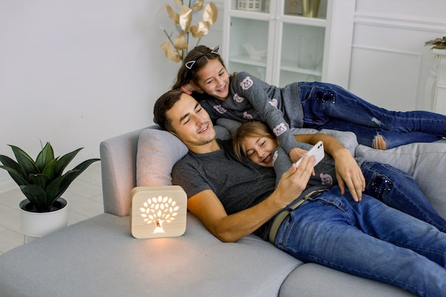 Happy family, young man with his two little sisters, having fun at cozy modern living room, lying on gray sofa with wooden hand made night lamp.