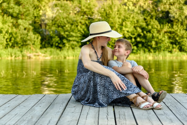 Happy family, young mother with her little son are sitting on the pier by the river