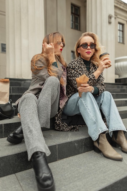 Happy Fanny young fashion girls in stylish sunglasses with fashionable casual clothes sitting on steps drinks coffee and eating profiteroles in the city Life is a oneway journey to be enjoyed