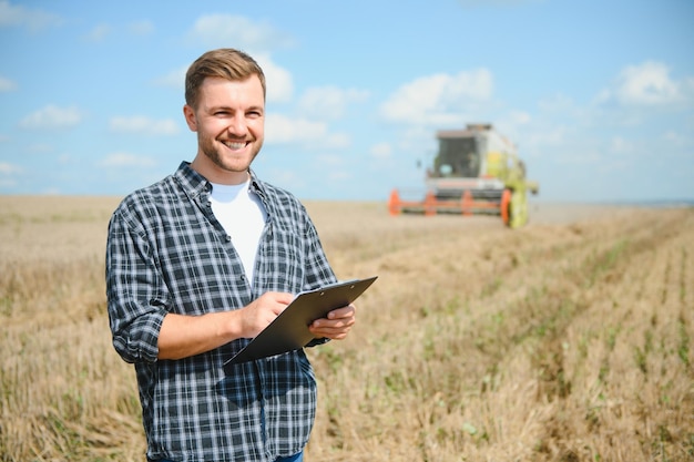 Happy farmer proudly standing in a field Combine harvester driver going to crop rich wheat harvest Agronomist wearing flannel shirt looking at camera on a farmland