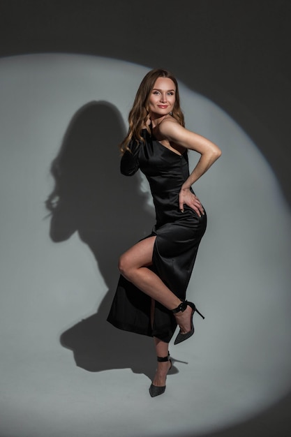 Happy fashion elegant pretty young woman model with slim sexy body in trendy stylish black evening dress posing and having fun in studio on dark background with round light