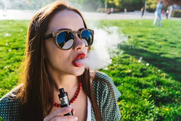 Счастливая мода улыбается женщина в солнцезащитных очках для некурящих vape на улице, дым