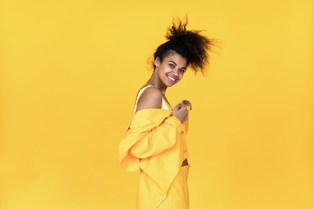 행복한 패션 젊은 아프리카 십대 소녀는 스타일리시한 노란색 슈트 옷을 입고 춤을 추며 배경에서 고립되어 웃는 혼혈 여성이 여름 컬러 스튜디오에 서서 카메라 초상화를보고 있습니다.
