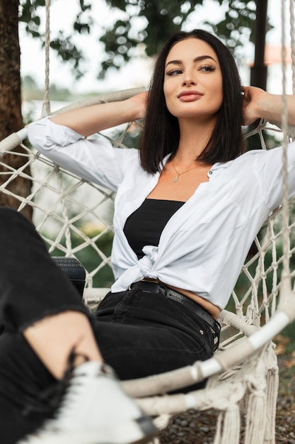 Happy fashionable beautiful business girl in stylish casual clothes with a white shirt and black jeans sits and rests in a knitted swing outdoors