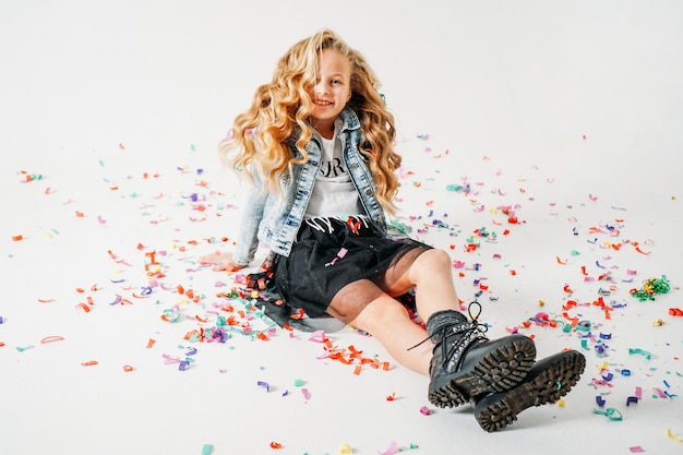 Happy fashionably dressed curly hair tween girl in in a denim jacket and black tutu skirt and rough boots sitting on white  with colorful confetti