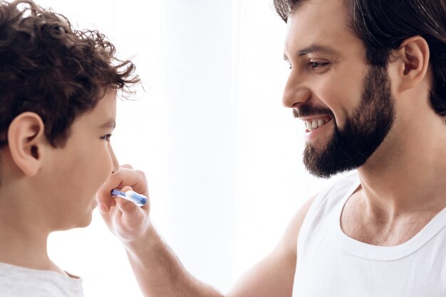 Happy father brushes teeth of young son with toothbrush.