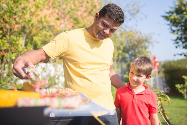 Happy father grilling meat and vegetables with son. Dark-haired man in yellow T-shirt putting sausage on BBQ grid. Smiling little boy standing next to him. BBQ, cooking, food, family concept