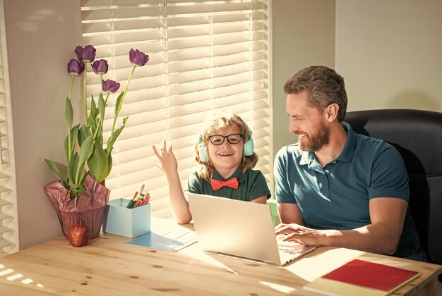 幸せな父は、ヘッドフォンで彼の学校の息子が学校に戻って自宅でラップトップで勉強するのを手伝っています