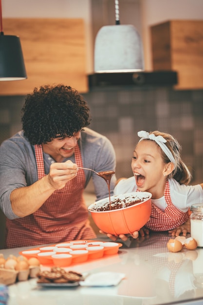 Happy father and his daughter are preparing cookies together in the kitchen. Father is pouring chocolate dough into molds and little girl wants to eat it.