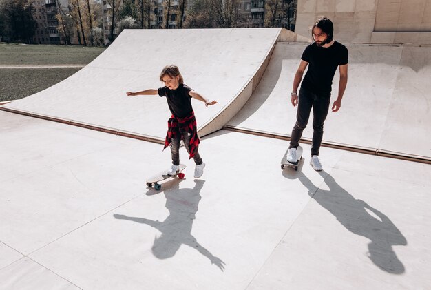 Happy father and his little son dressed in the casual clothes ride skateboards in a skate park with slides at the sunny day .