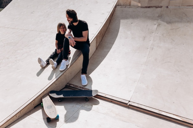 Il padre felice e suo figlio vestiti con abiti casual sono seduti e ridono dello scivolo in uno skate park accanto agli skateboard durante la giornata di sole.