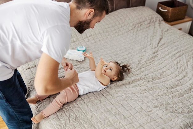 Happy father is putting clothes on his daughter while she is lying on a bed and talking to him