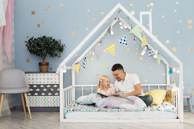Счастливый отец смеется со своей 3-летней девочкой, сидя на кровати Белого дома в детской спальне