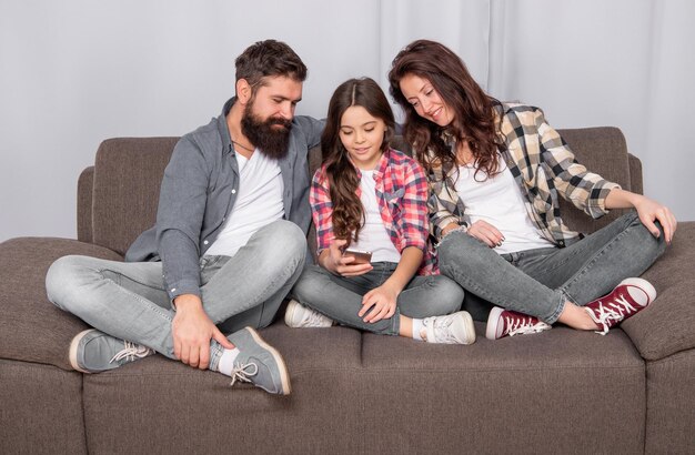 Happy father and mother look at girl child chatting in smartphone sitting on sofa chat