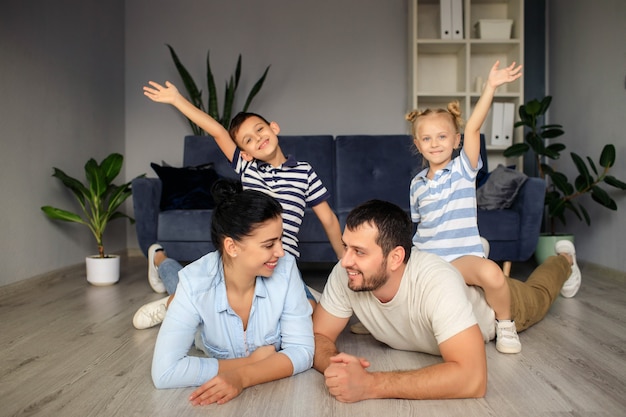 Happy father and mother with little children sitting on floor near sofa and laughing while having fun together at home during weekend