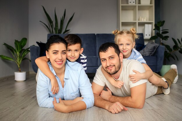 Happy father and mother with little children sitting on floor near sofa and laughing while having fun together at home during weekend