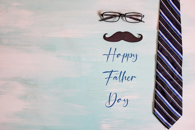 Happy Father's day concept. A black mustache paper, a blue necktie and glasses on wooden table.