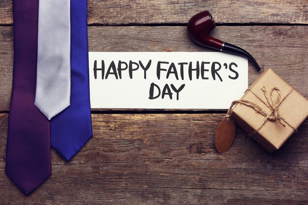 Happy Father's Day inscription with pipe and ties on wooden background Greetings and presents