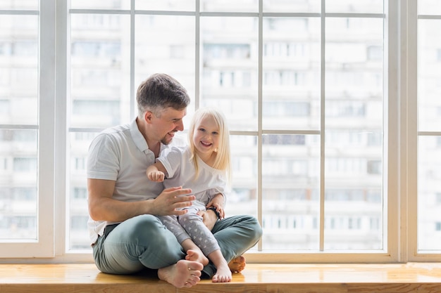 Happy father sitting on windowsill while giving a hug his years old daughter