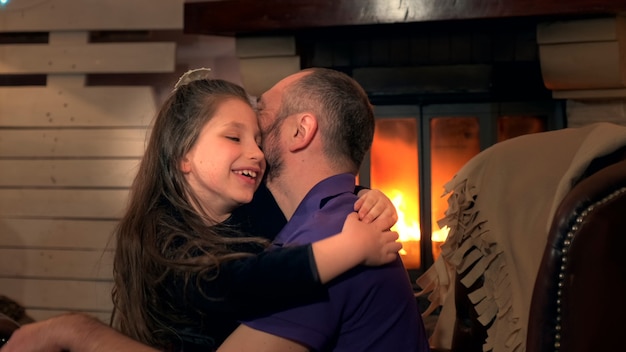 Счастливые отец и маленькая дочь обнимают друг друга в кресле у камина, которые они обожают ...