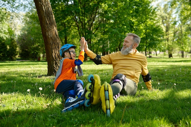 Happy father and son giving high five each other while rest on green grass meadow in city park after speed racing on rollerskates. Active family weekend