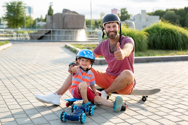 Happy father and son in helmets play in the park with a robot\
car that is controlled by a glove while sitting on\
skateboards.