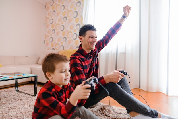 Photo happy father and son sitting on a floor and playing video games. both of them very excited. father is winning his son.