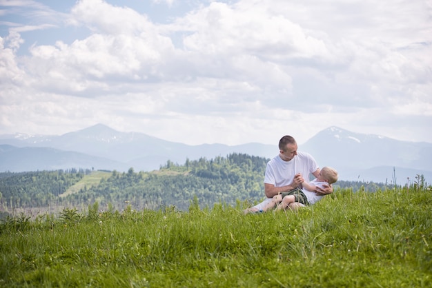 Happy father with his little son on his knees sits on meadow