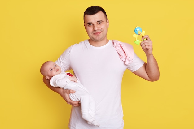 집에서 작은 아기 딸과 함께 행복 한 아버지, 노란색 벽 위에 절연 포즈를 취하는 동안 딸랑이 함께 돌보는 딸랑이 함께 젊은 아빠.