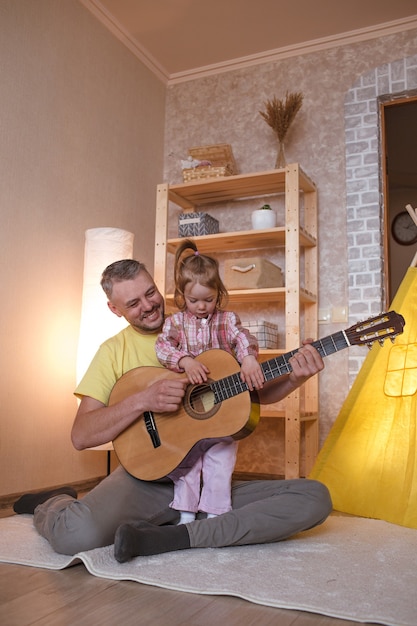 Un padre felice con una figlia piccola impara a suonare la chitarra seduto a casa sul pavimento vicino al teepee giallo. padre felice