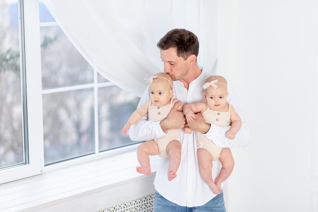 Happy father with twins daddy holds two newborn baby twin girls in a cotton suit at the window of the house hugging and kissing them fatherhood with two children