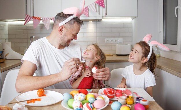 A happy father with two young daughters laughing and playing around having fun painting Easter eggs preparing for the Easter holiday