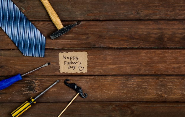 Happy Fathers Day gift box with tie, hammer, blue box and mustache on a rustic wood background.