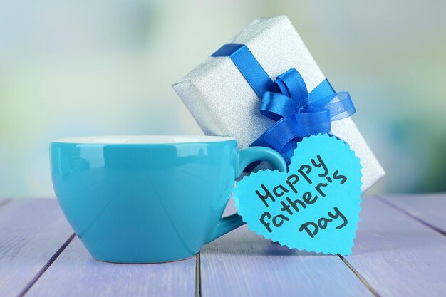 Happy Fathers Day tag with gift box and cup on wooden table on light background