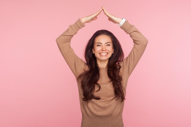 Happy to feel safe Portrait of cheerful young woman with brunette hair showing house roof symbol over head smiling joyfully gesture of protection insurance studio shot isolated on pink background