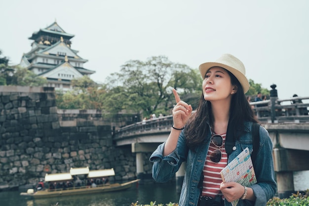 Happy female asian tourist holding guide book and paper map pointing at side on street. wood bamboo boar on river lake in background under stone bridge. osaka castle surrounding by strong wall japan.